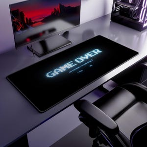 Puede incluir: Una alfombrilla de escritorio rectangular negra con un gráfico blanco pixelado que dice GAME OVER, seguido del texto DO YOU WANT TO CONTINUE? y las opciones NO y YES con una flecha de cursor apuntando a la palabra YES en estilo de videojuego retro.