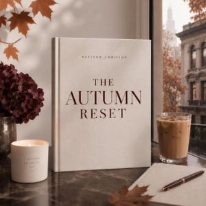 Może przedstawiać: Książka w twardej oprawie zatytułowana The Autumn Reset autorstwa Refined Ambition z kremową okładką, umieszczona obok białej świecy z tekstem, szklanki mrożonej kawy, notatnika i długopisu, na tle okna z widokiem na jesienne drzewa.