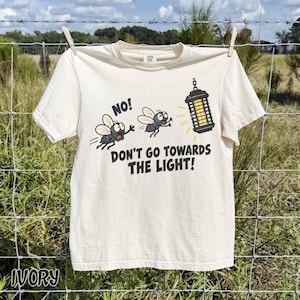 May include: An ivory short-sleeve t-shirt featuring a graphic of two cartoon flies and a bug zapper. The text reads, NO! DON'T GO TOWARDS THE LIGHT! The flies appear to be warning each other as they fly toward the glowing black and yellow bug zapper.
