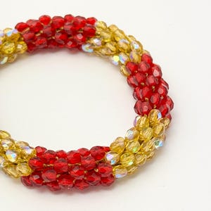 May include: A circular bracelet constructed from small, faceted beads in alternating segments of vibrant red and translucent gold. The beads are arranged in a dense, multi-row pattern, creating a textured, cylindrical band with a reflective finish.