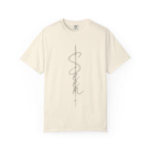 May include: A cream-coloured short-sleeve t-shirt featuring a vertical graphic design in the centre. The design consists of the word "Seen" written in a cursive script font, intertwined with a thin, vertical needle-like line and a small star.