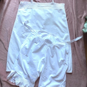 May include: A white cotton camisole featuring delicate lace trim along the neckline and thin shoulder straps, paired with matching white cotton bloomers that have an elasticized waist and decorative eyelet lace ruffles at the leg openings.
