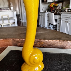 May include: A tall, bright yellow ceramic vase with a smooth, glossy finish. The base features a distinct, rounded spiral or snail-like shape that transitions into a long, slender, slightly curved neck that flares open at the top.