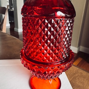 May include: A vibrant red glass candy dish featuring a diamond-cut pattern across the cylindrical body and the flared base. The piece includes a matching domed lid with a faceted top, showcasing a translucent finish that catches the light.