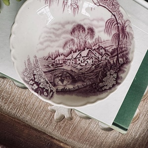 May include: A round ceramic bowl with a scalloped edge featuring a dark purple transferware print of a rural landscape. The scene depicts a stone bridge over a stream, several cottages, and willow trees against a white background.
