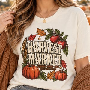 May include: A cream-colored t-shirt featuring a graphic print with the text HARVEST MARKET, PUMPKINS, APPLES, CIDER, and OPEN SEPTEMBER-NOVEMBER. The design includes illustrations of two orange pumpkins, two red apples on a branch, wheat stalks, and autumn leaves.