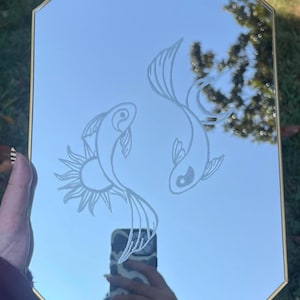 May include: A rectangular wall mirror with a thin gold-toned metal frame. The glass surface features a white etched design of two koi fish swimming in a circular motion, with one fish positioned near a sun-like graphic element.