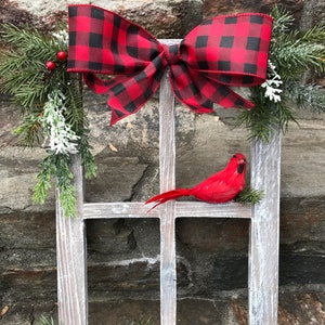 May include: A rustic wooden window frame decorated for the holidays. A large red and black buffalo plaid bow sits at the top. A red cardinal bird rests on a window pane. Greenery, berries, and a pinecone complete the festive arrangement.