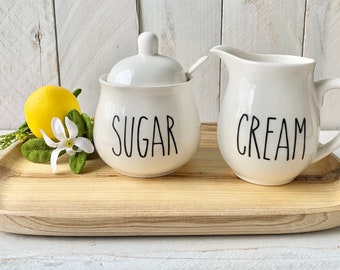 Puede incluir: Juego de azúcar y crema de cerámica blanca con letras negras. El azucarero tiene una tapa y una cucharita pequeña. La jarra de crema tiene un asa. Ambos recipientes dicen "SUGAR" y "CREAM" respectivamente. El juego está en una bandeja de madera.