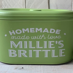 May include: A green oval-shaped container with a white lid. The container has the text "HOMEMADE. made with love MILLIE'S BRITTLE" printed on the front.
