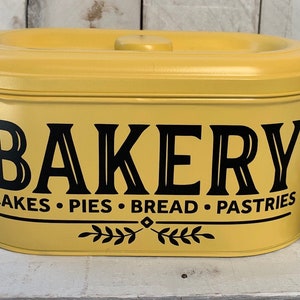 May include: A yellow metal bakery storage container with a black inscription that reads "BAKERY Cakes • Pies • Bread • Pastries".