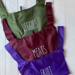 May include: Three reusable shopping bags with different labels. The top bag is green and says "VEGGIES". The middle bag is burgundy and says "MEATS". The bottom bag is purple and says "FRUIT".