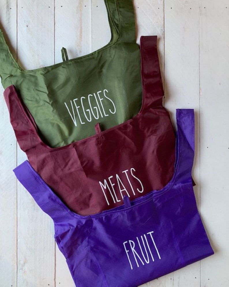 May include: Three reusable shopping bags with different labels. The top bag is green and says "VEGGIES". The middle bag is burgundy and says "MEATS". The bottom bag is purple and says "FRUIT".