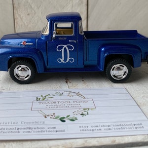May include: A blue toy pickup truck with a white monogram "D" on the side. The truck has a chrome bumper and wheels. The truck is sitting on a wooden surface.