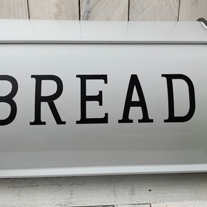 May include: White bread box with the word "BREAD" in large black letters. The box has a curved top and black trim. A green wreath with a cream-colored ribbon is on the right side.