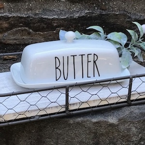 May include: White ceramic butter dish with the word "BUTTER" printed in black on the lid. The dish is sitting on a white wooden tray with a wire mesh frame.