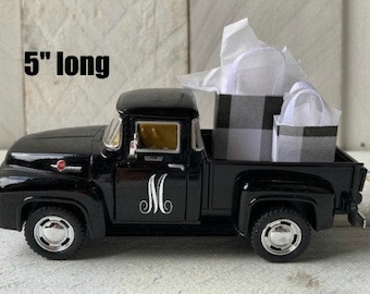 May include: A black vintage-style toy truck, 5 inches long, with the letter "M" on the door. The truck bed holds two small gift boxes, one with a black and white plaid pattern and the other with a white handle.