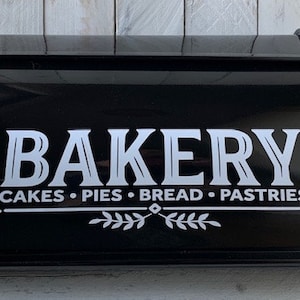 May include: Black bread box with a curved top and a roll-top lid. The word "BAKERY" is displayed in white, stylized lettering, along with the words "CAKES • PIES • BREAD • PASTRIES". A decorative leaf design is below the text.
