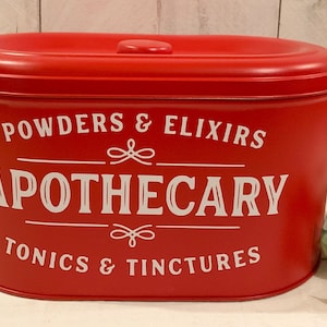 May include: Red metal canister with a white lid and white lettering that reads "POWDERS & ELIXIRS APOTHECARY TONICS & TINCTURES".