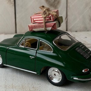 May include: A green toy car with a red and white striped gift box on top. The car is a classic model with a rounded body and a chrome bumper.