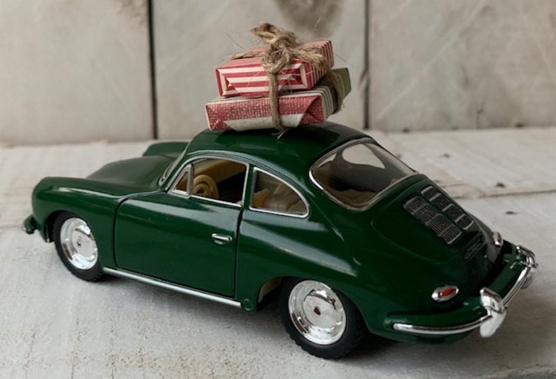 May include: A green toy car with a red and white striped gift box on top. The car is a classic model with a rounded body and a chrome bumper.