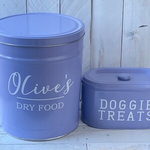 May include: Two purple metal canisters with white lettering. The larger canister says "Olive's Dry Food" and the smaller canister says "Doggie Treats".