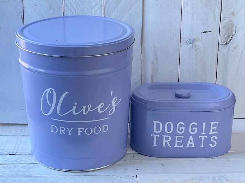 May include: Two purple metal canisters with white lettering. The larger canister says "Olive's Dry Food" and the smaller canister says "Doggie Treats".