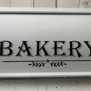 May include: White metal bread box with a black inscription that reads "BAKERY" with a decorative flourish of leaves below the word.