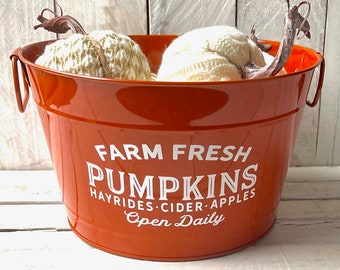 May include: Orange metal bucket with white lettering that reads "Farm Fresh Pumpkins Hayrides Cider Apples Open Daily". The bucket is filled with two white knitted pumpkins.