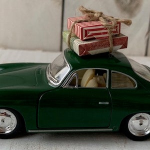 May include: A green toy car with a red and white striped gift box on top. The car is a classic model with a rounded body and chrome wheels.