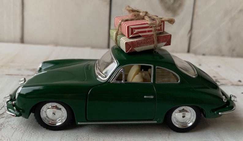 May include: A green toy car with a red and white striped gift box on top. The car is a classic model with a rounded body and chrome wheels.