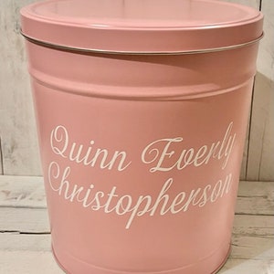 May include: A pink metal storage container with a white lid. The container has the name "Quinn Everly Christopherson" written on the side in white lettering.