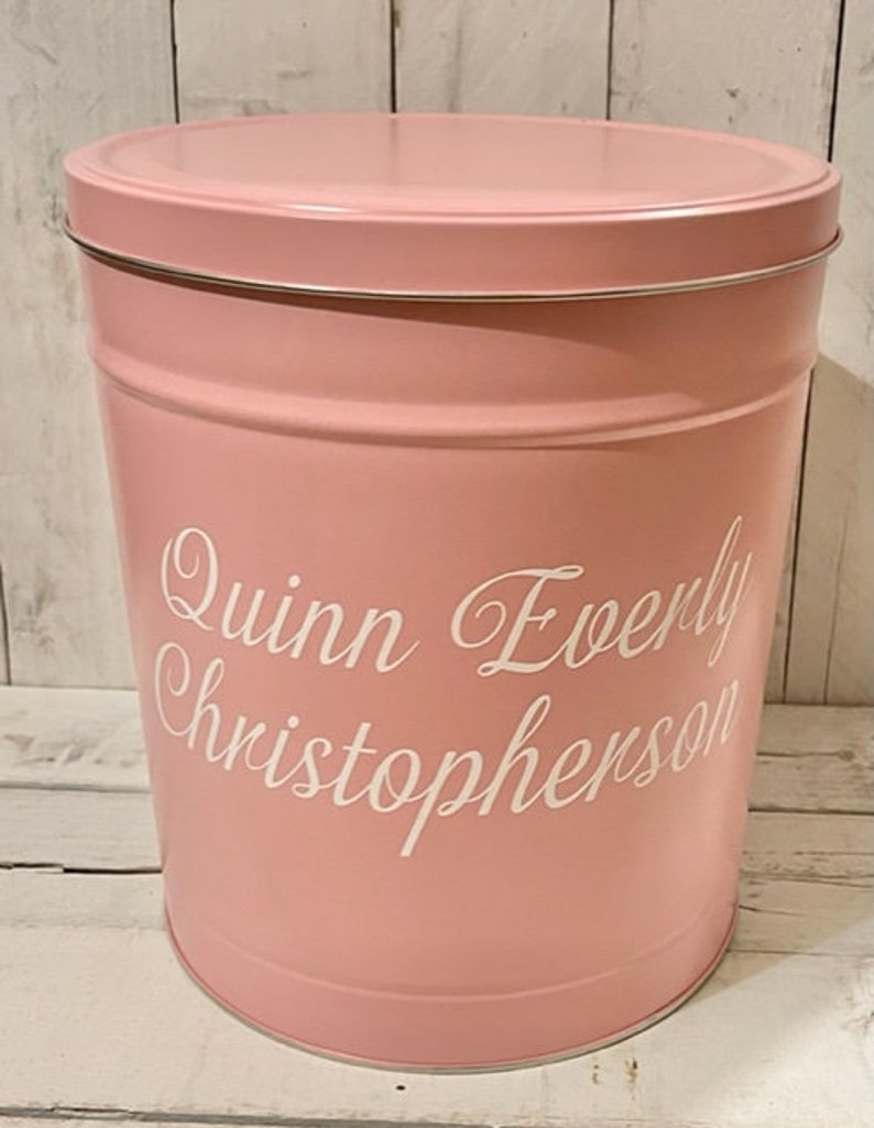 May include: A pink metal storage container with a white lid. The container has the name "Quinn Everly Christopherson" written on the side in white lettering.