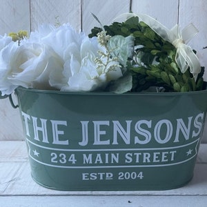 May include: A green metal planter with white lettering that reads "The Jensons 234 Main Street Estd 2004". The planter is filled with white flowers and a green wreath.