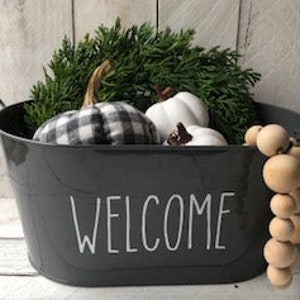 May include: A gray metal container with black handles and the word "WELCOME" printed in white. The container is filled with a green wreath, white pumpkins, and a black and white checkered pumpkin. A wooden bead garland is on the right side of the container.