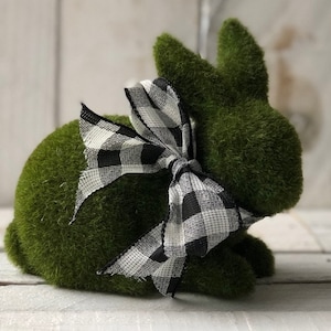May include: A green, moss-covered bunny figurine with a black and white checkered bow. The bunny has upright ears and is posed in a seated position. This decorative item is suitable for Easter or spring decor.