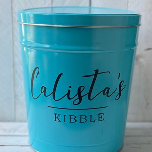 May include: A turquoise blue metal container with a silver lid. The container has the text "Calista's Kibble" printed on the front.