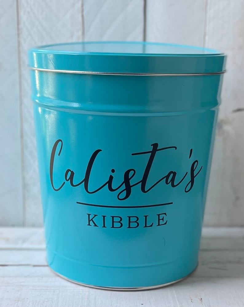 May include: A turquoise blue metal container with a silver lid. The container has the text "Calista's Kibble" printed on the front.