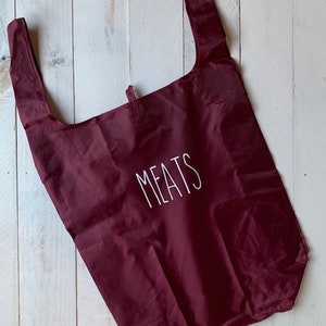 May include: A burgundy reusable shopping bag with the word "MEATS" printed in white on the front.