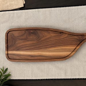 May include: A rectangular dark brown wooden serving board with a smooth finish and visible natural wood grain patterns. The board features a rounded handle with a circular cutout at one end and a shallow carved groove along the perimeter.