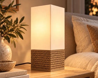 May include: A square table lamp with a white fabric shade and a tan woven cane base. The lamp emits a warm glow. Text on the image reads: Warmth in every corner, Cozy Nook, Tan. The lamp features a clean, geometric design with a textured base.