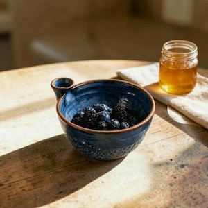 Peut inclure: Un bol en céramique bleu profond avec une petite poignée latérale rempli de mûres et de myrtilles fraîches. À côté du bol se trouve un petit pot en verre transparent rempli de miel doré sur une serviette en tissu clair.
