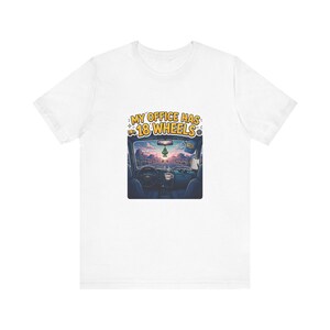 May include: A white t-shirt featuring a graphic of a truck interior looking out onto a desert highway at sunset. The text above the image reads MY OFFICE HAS 18 WHEELS in yellow letters. The scene includes a steering wheel and air freshener.