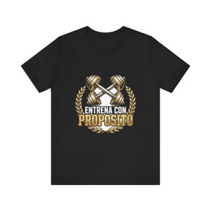 May include: A black short-sleeve t-shirt featuring a graphic of two crossed gold dumbbells centered within a gold laurel wreath. Below the dumbbells, the text "ENTRENA CON PROPOSITO" is printed in bold, gold, capitalized lettering.