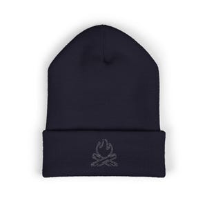 May include: A dark navy blue cuffed beanie made of knit fabric. The front of the cuff features a small, white, embroidered line-art graphic of a campfire with two crossed logs and flames rising above them, centered on the folded brim.