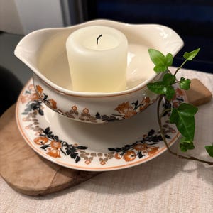 May include: A cream-colored ceramic gravy boat with a scalloped rim and a floral pattern in orange and black, resting on a matching saucer. A white pillar candle sits inside the gravy boat, and a green ivy vine is draped over the side.