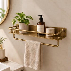 Pode incluir: Uma prateleira de casa de banho de parede em latão com um corrimão e uma barra inferior para toalhas. A prateleira contém uma planta verde num vaso claro, uma garrafa de vidro âmbar com o rótulo Hand Wash e um pequeno recipiente.