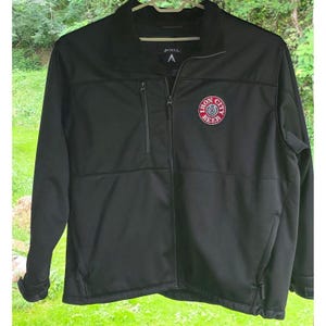 May include: A black full-zip jacket featuring a mock neck collar and long sleeves. The chest displays a circular red and white patch with the text Iron City Beer. It includes a vertical zippered pocket on the right chest and two side pockets.