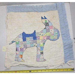 May include: A square quilted fabric panel featuring a patchwork dog appliqué in various pastel colors. The panel has a light blue border and a white background. A yellow measuring tape at the bottom shows the width is approximately 15 inches.