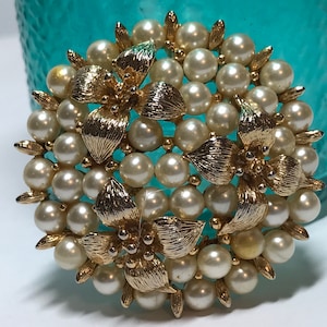 May include: A vintage brooch featuring a circular design of faux pearls and gold-toned metal floral accents. The brooch has a cluster of off-white pearls and gold-colored leaves and petals. The brooch is a classic piece of jewelry.
